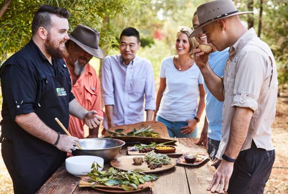 北領地（Northern Territory）尤拉拉（Yulara）「叢林食物」體驗（Bush Food Experience）的參加者圍站在擺滿叢林食物的戶外餐桌前©Voyages Indigenous Tourism