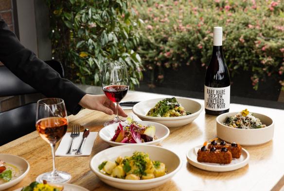 維多利亞（Victoria）摩寧頓半島（Mornington Peninsula）Jackalope Rare Hare Wine &amp; Food Store的餐桌上擺滿新鮮美食，一人正伸手拿桌上的紅酒©Amy Whitfield