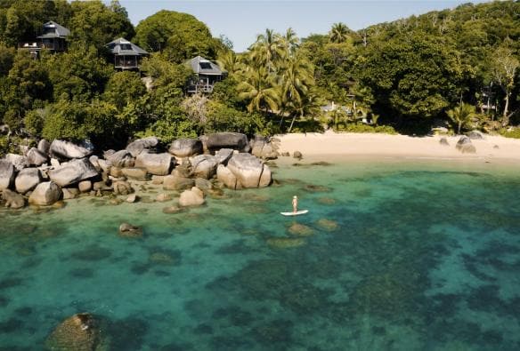 空中俯瞰昆士蘭州大堡礁班達拉島海岸附近清澈碧藍的水面、幾堆巨石、白色沙灘和綠色棕櫚樹©澳洲旅遊局