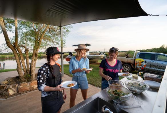 三名參加了北領地（Northern Territory）貝利泉（Berry Springs）Barramundi Adventures Darwin體驗的團友正享用自助餐，身後是一條水道和一艘船©澳洲旅遊局
