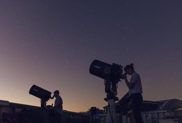 兩個人在夜幕初垂時用昆士蘭沙勒維爾宇宙中心的望遠鏡仰望星空，地平線上可以看到夕陽的霞光©昆士蘭旅遊及活動推廣局