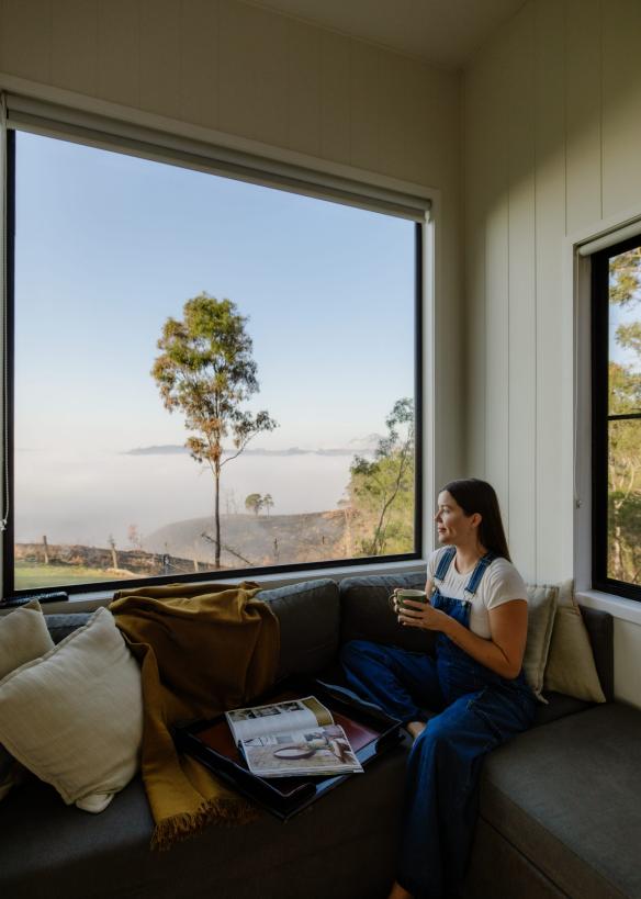 一個人坐在昆士蘭美景環區Baya Tiny House內的長椅上透過窗戶欣賞後山腹地景致©澳洲昆士蘭州旅遊及活動推廣局